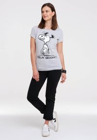 T-shirt gris avec un graphique noir représentant un chien de dessin animé et le texte "FEELIN' GROOVY!" Un pantalon noir et des baskets blanches complètent la tenue.
