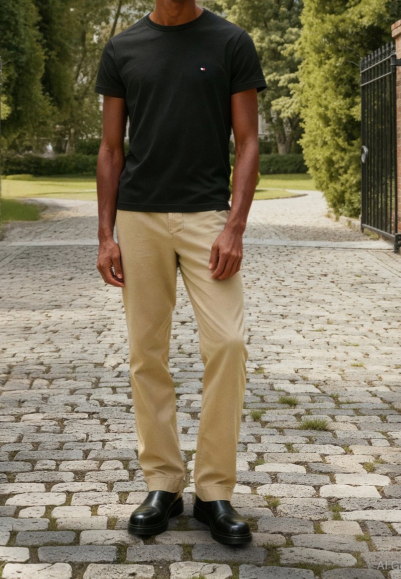 Homme debout sur un chemin pavé portant un t-shirt noir, un pantalon beige et des chaussures noires, avec de la verdure et une porte en arrière-plan.