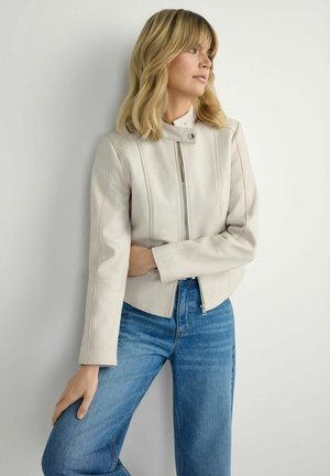 Kobieta o blond włosach do ramion, ubrana w kremową kurtkę na zamek i niebieskie dżinsy, stojąca na tle jednolitej jasnej ściany.