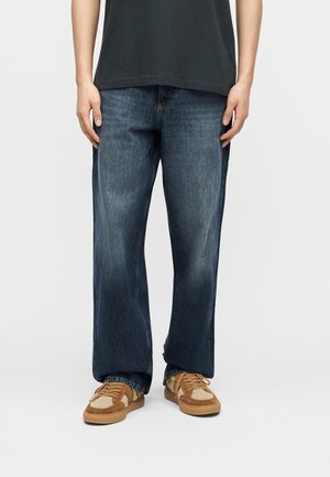 Person trägt dunkelblaue lockere Jeans und braune Sneaker mit beigen Akzenten, steht vor einem schlichten hellen Hintergrund.