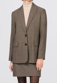 Veste blazer marron à carreaux avec revers crantés, deux boutons et poches avant, portée sur un col roulé beige clair et une jupe assortie.