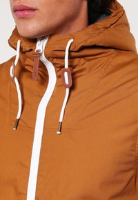 Brun hættejakke med hvid lynlås, der har justerbare snore, metalliske brune detaljer og en glat, let stoftekstur.