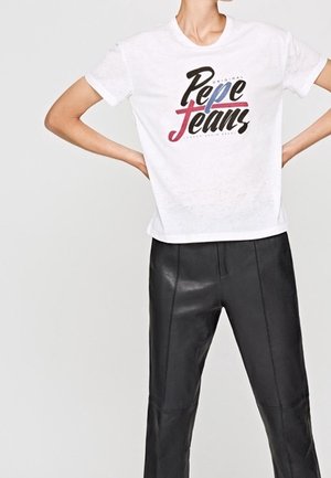 Persoon die een wit Pepe Jeans t-shirt en zwarte leren broek draagt, staand met handen in de zij tegen een effen lichte achtergrond.