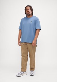 T-shirt en coton bleu clair avec un petit logo jaune sur la poitrine, associé à un pantalon beige et des chaussures de sport grises avec des accents blancs.