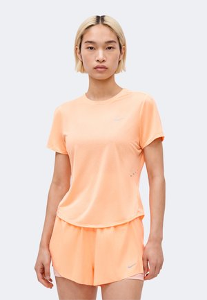Kobieta stoi przodem, ubrana w pasujący jasnopomarańczowy zestaw Nike z krótkim rękawem i szortami.