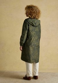 Moteris su garbanotais plaukais, vilkinti alyvuogių žalumos gėlių rašto su kapučinu lietpaltį ir baltas kelnes, stovi šonu prieš kreminę fono.