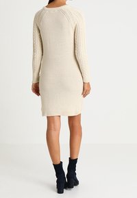 Pletené šaty v krémové barvě, s texturovaným vzorem a dlouhými rukávy. Stylizováno s námořnickými kotníkovými botami, sahajícími do poloviny stehna. Hladký výstřih a lem.