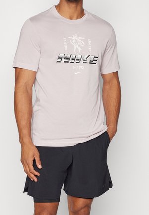 Homme portant un t-shirt Nike rose clair avec un texte graphique et un short de sport noir, debout avec une main dans la poche.