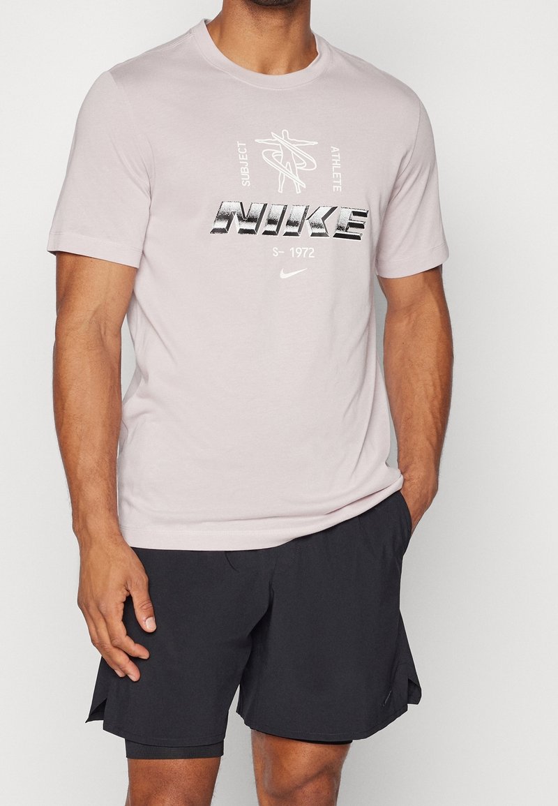 Homme portant un t-shirt Nike rose clair avec un texte graphique et un short de sport noir, debout avec une main dans la poche.