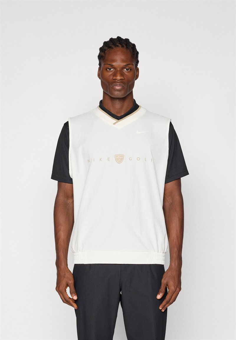 White Nike Golf vest over a black shirt, featuring a v-neck, ribbed hem, and logo details in gold. Smooth texture, sleeveless design.