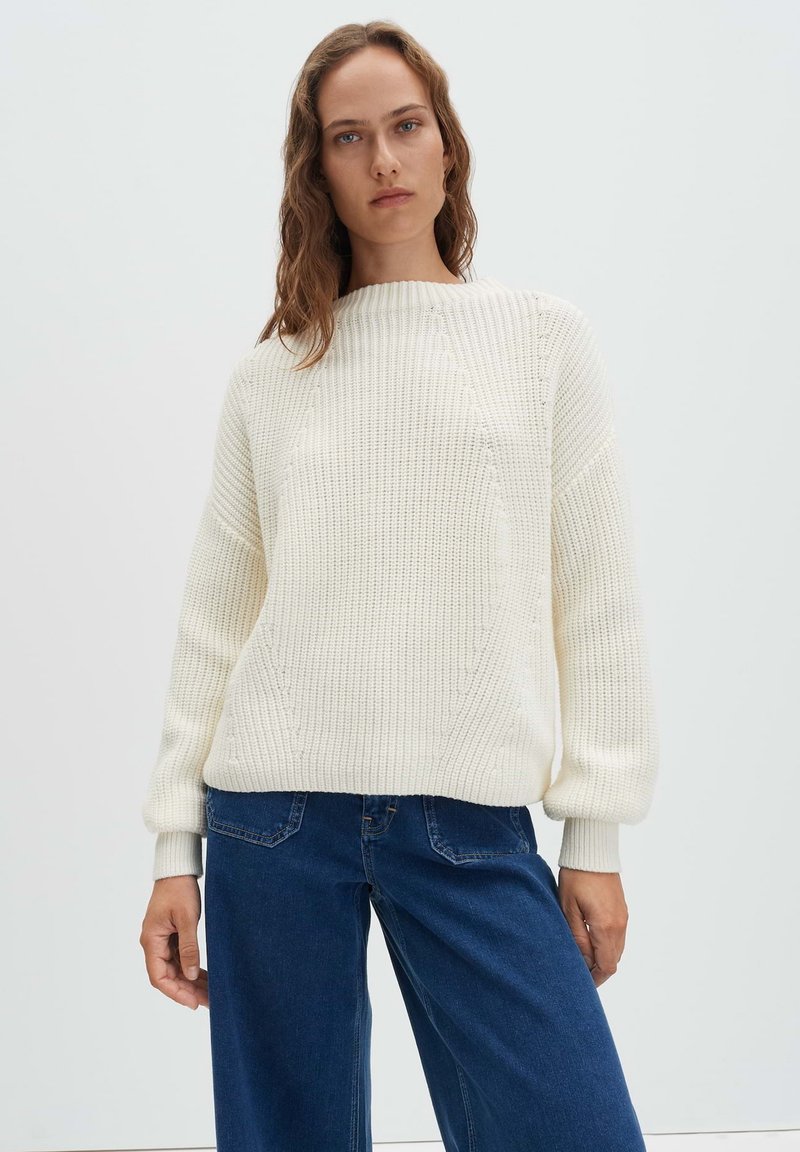 Pull blanc en maille avec un motif texturé, doté d'un large col et de poignets côtelés, associé à un jean à jambes larges bleu foncé.