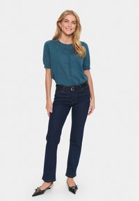 Žalias megztinis su trumpomis rankovėmis, su mygtukų detale, derinamas su tamsiai mėlynomis džinsomis ir juodais batais su smailiu priekiniu odeliu ir dekoratyvinėmis detalėmis.