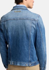 Mężczyzna w niebieskiej kurtce dżinsowej i beżowych spodniach, pokazany od tyłu na białym tle.