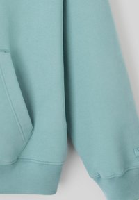 Gros plan d'une manche et d'une poche de sweat-shirt bleu clair avec des détails de couture en côte sur un fond neutre.