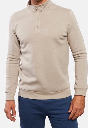 Mand iført en beige pullover med kvart-lynlås, ribstrikkede manchetter og kant, kombineret med blå bukser.