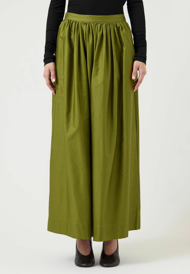 Naine, kes kannab kõrge vöökohaga, laia säärega oliivrohelisi pükse, millel on kogutud talje ning musta pika varrukaga topi, seisab mustades kingades.