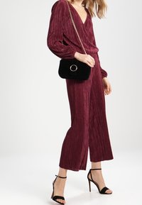 Burgundy pleated jumpsuit with V-neck, long sleeves, and wide legs, paired with a black quilted handbag and strappy black heels.