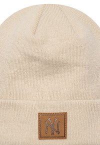Beanie beige in maglia con risvolto, caratterizzato da una toppa in pelle marrone rettangolare stampata con un logo stilizzato. Texture morbida.