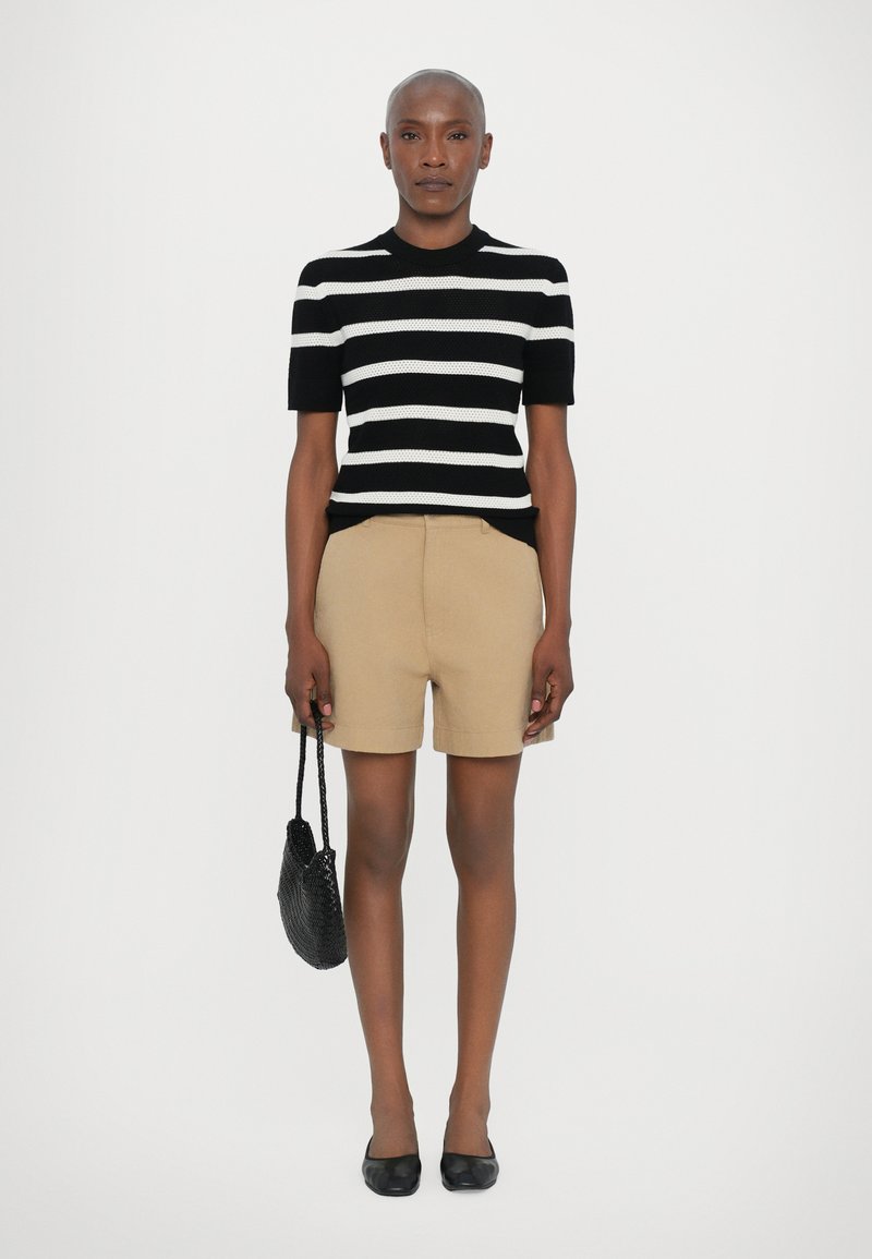 Haut en tricot à rayures noir et blanc à manches courtes, associé à un short beige cintré. Tient un sac à main tissé noir et porte des ballerines noires.