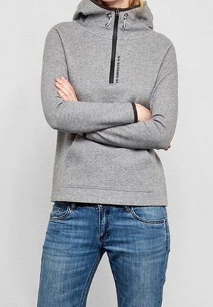 Personne portant un sweat à capuche gris clair avec une fermeture éclair noire et un jean bleu, bras croisés, debout devant un fond uni.
