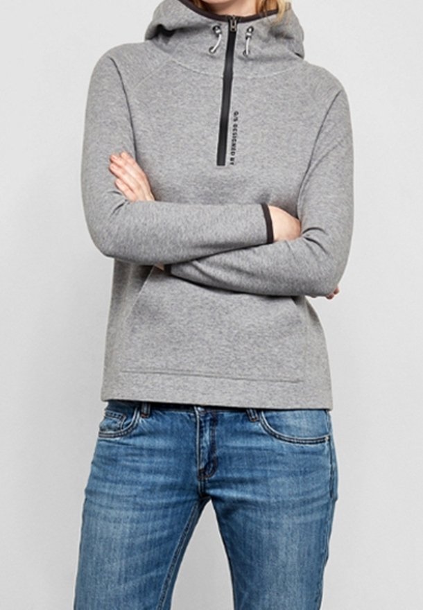 Personne portant un sweat à capuche gris clair avec une fermeture éclair noire et un jean bleu, bras croisés, debout devant un fond uni.
