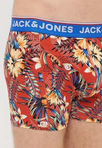 Red floral-patterned boxer briefs with a blue elastic waistband featuring "JACK & JONES" logo. Material appears stretchy with a smooth texture.