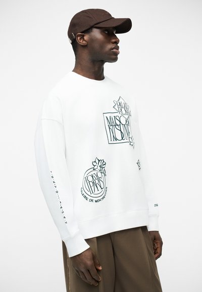 Άνδρας που φοράει καφέ καπέλο, λευκή φούτερ με μαύρο γραφικό κείμενο και καφέ παντελόνι, στραμμένος πλαγίως προς λευκό φόντο.