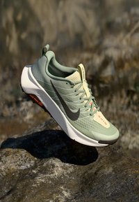 O sapatinho de corrida verde da Nike possui um cabedal texturizado, entressola branca, logotipo Swoosh preto e detalhes laranja na lingueta e na sola exterior.