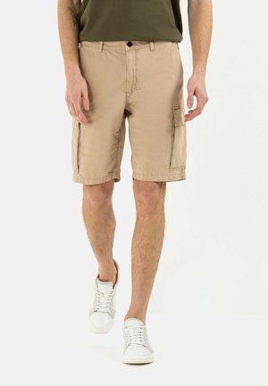 Hombre con pantalones cortos cargo beige y zapatillas blancas caminando hacia adelante sobre fondo blanco.