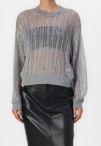 Srebrny metaliczny sweter dzianinowy z szarym prążkowanym kołnierzem i mankietami, z przejrzystym wzorem i czarnym blokiem dekoracyjnym na wysokości klatki piersiowej. W połączeniu z czarną skórzaną spódnicą.