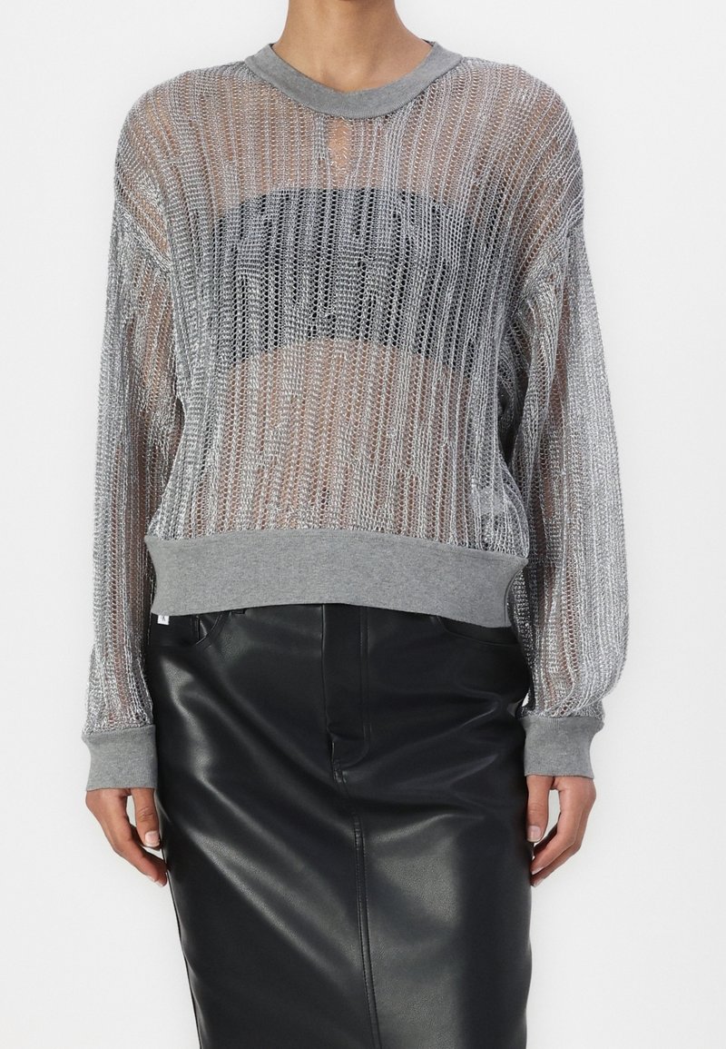 Srebrny metaliczny sweter dzianinowy z szarym prążkowanym kołnierzem i mankietami, z przejrzystym wzorem i czarnym blokiem dekoracyjnym na wysokości klatki piersiowej. W połączeniu z czarną skórzaną spódnicą.