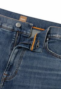 Mörkblå denimjeans med dragkedja och knappstängning, orange sömnadsdetaljer och en brun märke på midjebandet.