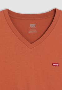 Rozsdabarna v-nyakú pamut póló. Bordás gallérral rendelkezik, és egy kis piros Levi’s logó található a bal mellrészen. Simás textúrájú.