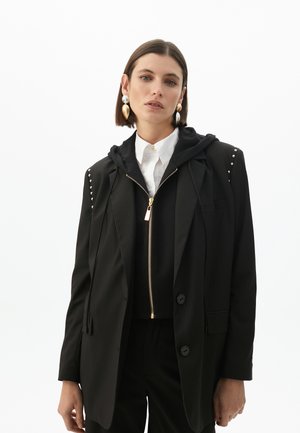 Blazer nero a righe con cappuccio rimovibile nero, zip dorata e decorazioni argentate sulle spalle. Indossato su una camicia bianca e pantaloni abbinati.
