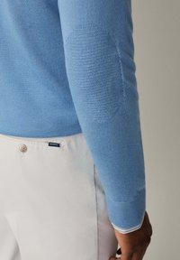 Blaues Sweatshirt mit strukturiertem Ellbogenpatch, kombiniert mit hellbeigen Hosen, die einen Knopf und ein Markenetikett aufweisen.