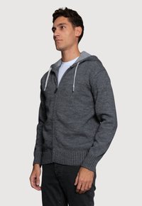 Szara bluza zapinana na zamek z kontrastującym jasnoszarym kapturem, z prążkowanymi mankietami i dołem, z teksturowanej dzianiny, noszona na białym t-shircie.