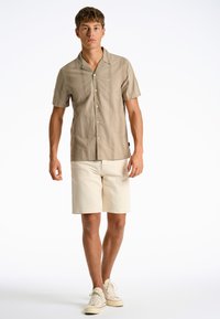 Korte mouwen overhemd in lichtbeige met verticale patronen, gecombineerd met crèmekleurige shorts en beige sneakers met rode accenten.