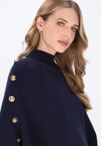 Pulover bleu închis cu gât înalt, mâneci lungi și nasturi decorativi aurii pe umeri. Textură moale, cu o croială lejeră.