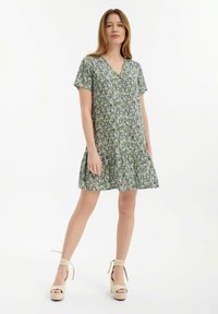Grünes Blumenkleid mit kurzen Ärmeln, V-Ausschnitt, lockerer Passform und ausgestelltem Rock; kombiniert mit hellbeigen Absatzsandalen.