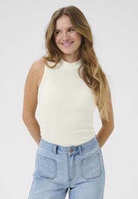 Top senza maniche di color crema leggero con collo alto e texture a costine, abbinato a jeans azzurri a vita alta con tasche applicate.