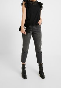 Top nero texture con maniche a volant, abbinato a jeans grigi a vita alta e stivaletti neri, mentre tiene una pochette nera con decorazioni.