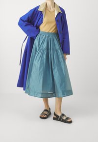 Cappotto esterno blu con colletto beige, top beige, gonna a strati turchese e sandali neri con strisce bianche. Texture morbide e vestibilità rilassata.
