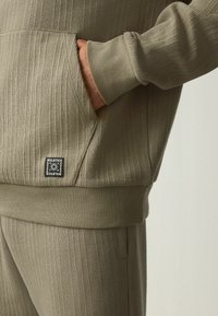 Sudadera de canalé en color caqui con bolsillo lateral y parche de marca. Textura suave, corte holgado y mangas con puños.
