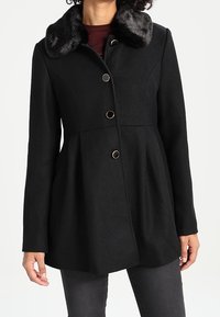 Cappotto nero con tessuto texturizzato, dotato di un colletto in pelliccia sintetica, bottoni in tono oro e silhouette svasata; maniche lunghe e taglio fino ai fianchi.