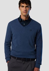 Dunkelblauer V-Ausschnitt-Pullover aus weichem Material, mit einem Logo auf der Brust und gerippten Bündchen. Über einem Hemd mit Kragen getragen.