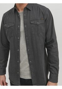 Camisa de mezclilla gris con mangas largas, cierre frontal de botones, dos bolsillos en el pecho y un acabado texturizado. Se lleva sobre una camiseta gris claro.