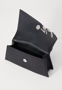 Bolso de mano de cuero negro con un diseño abierto, que cuenta con un acento de logo plateado y un cierre magnético en la parte frontal. Textura suave sin patrones.