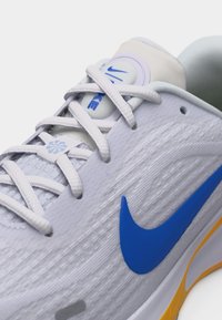 Hvit Nike løpesko med blå swoosh, hvite skolisser og gul detalj på sålen, sett i nærbilde ved skolissene og sidepanelet.