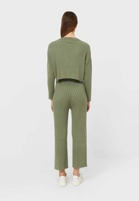 Olijfgroene geribbelde gebreide twee-delige set, bestaande uit een cropped top met lange mouwen en hoog- taille rechte broek. Witte sneakers.