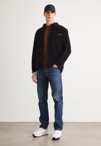 Čierna kordová košela-jacket cez hnedý rebrovaný sveter, v kombinácii s modrými džínsami a bielymi teniskami. Model nosí čiernu čiapku.